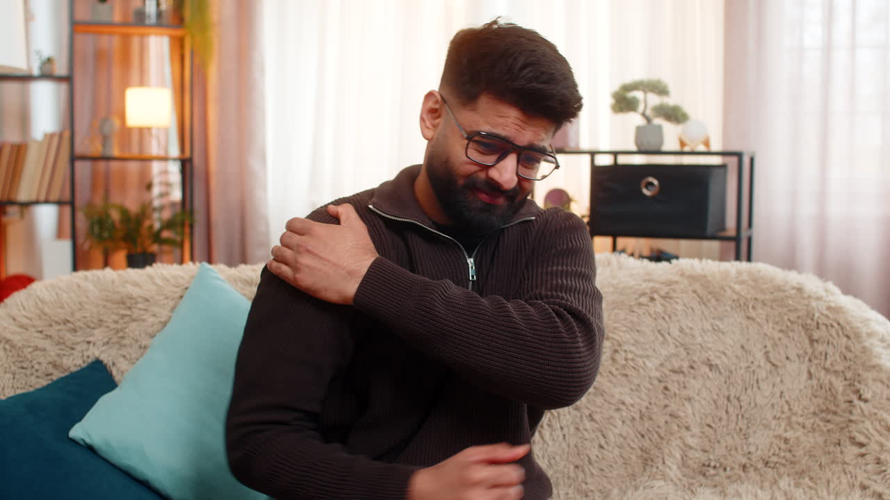 Indian man sitting at home holds shoulder with discomfort reacting to physical pain and tension