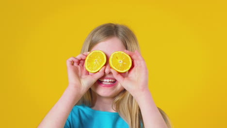 studio portrait of girl holding two orange halves in front of eyes against yellow background