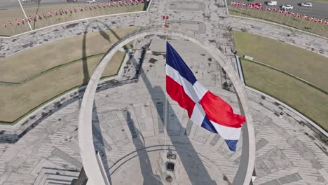 Toma-Aérea-De-Arriba-Hacia-Abajo-De-La-Bandera-Que-Se-Balancea-En-El-Famoso-Monumento-En-La-Plaza-De-La-Bandera,-República-Dominicana