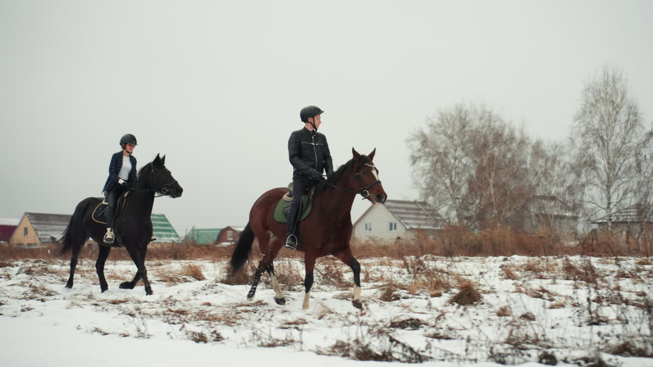 Due cavalieri a cavallo in un campo innevato, istruttori locali durante l'esercizio mattutino vicino alle case del villaggio, cavalli marroni e neri, selle in cuoio, caschi, erba gelata, cielo coperto, toni tenui cinematografici.