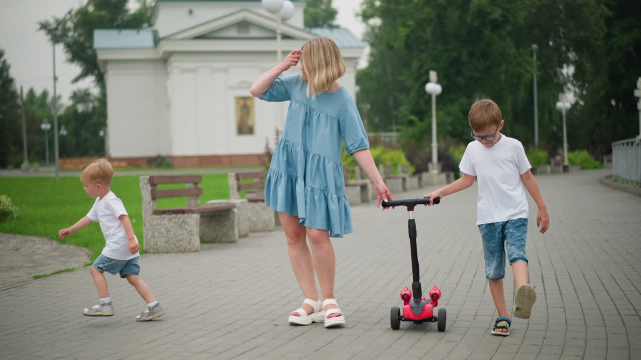una madre camina de la mano con su hijo menor mientras sostiene un scooter junto con su hijo mayor, el niño menor, distraído por algo, comienza a correr hacia la dirección