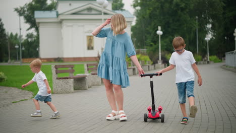 a mother walks hand-in-hand with her younger son while holding a scooter along with her older son, the younger child, distracted by something, starts running toward the direction