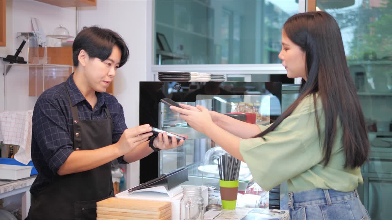 Asian woman customer and barista using a smart phone to scan QR code tag on another smartphone in a coffee shop or restaurant to accepted generate digital pay without money.