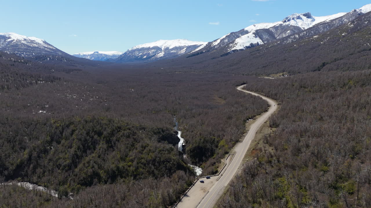 Drone captures Cascada Vuliñanco waterfall, curvy road into horizon, surrounded by forest, snow-capped mountains background - Neuquen, Argentina