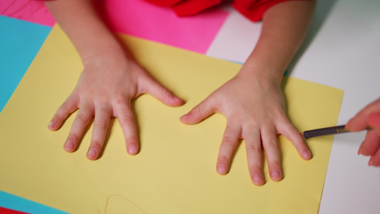 Little kid's hands on the yellow sheet of paper. Mother drawing a line around baby fingers. Close up.