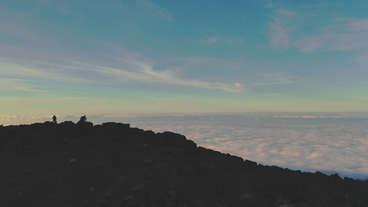 Three hikers climbing on a cliff below the Pico de Teide mountain on Canary Islands at sunset, watching a dense cloud inversion below the hillside. Hikers watching a cloud inversion at sunset 4K.