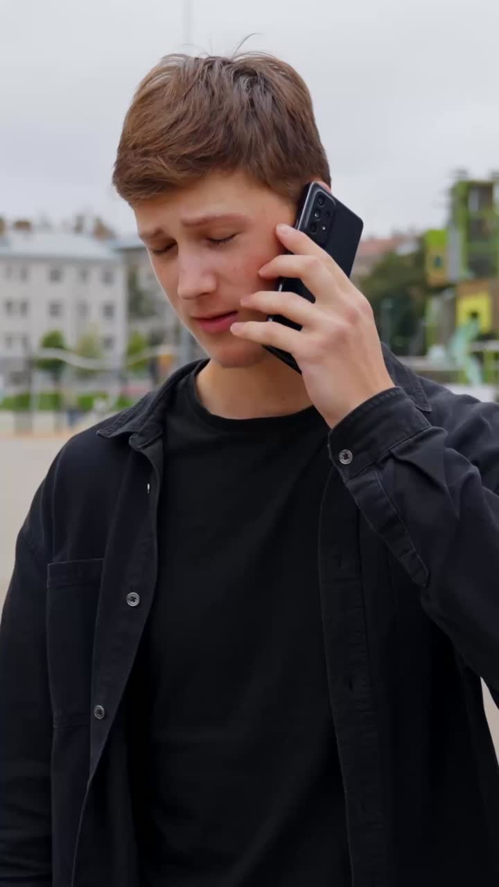 Man in dark clothing talks on phone with serious facial expression while standing outdoors