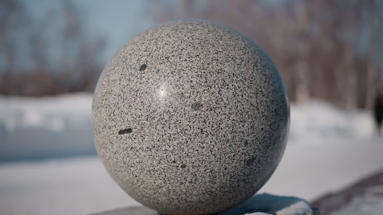 circular granite sphere on snow covered pedestal with blurred snowy park background under crisp winter daylight showcasing textured stone surface and focus winter environment