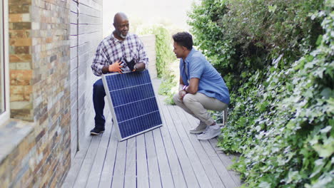 african american father showing solar panel to adult son in sunny garden, copy space, slow motion