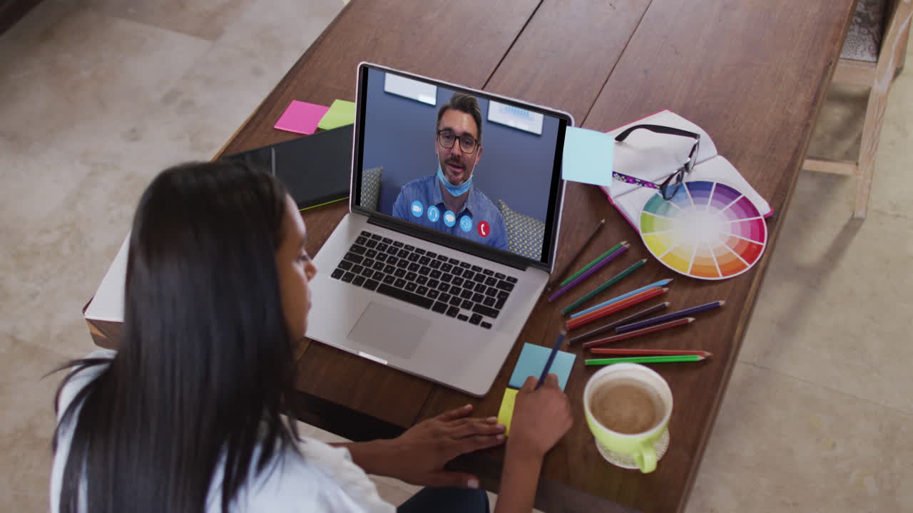 Mujer caucásica usando una computadora portátil en una videollamada con un colega y tomando notas.