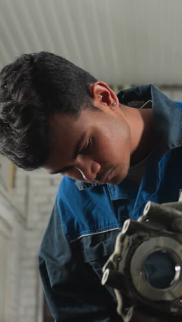mecánico en uniforme azul inspeccionando componentes del motor en un taller mecánico con vista parcial de otra persona que lo observa, centrado en el trabajo de reparación y la maquinaria