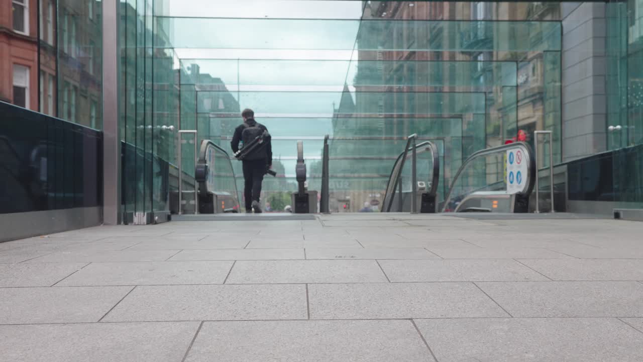 Man carrying camera and tripod enters large glass building with escalators