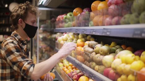 giovane in camicia a quadri, con maschera nera e guanti di plastica trasparenti, fa la spesa di frutta al supermercato durante la pandemia di quarantena del coronavirus covid-19. vista laterale