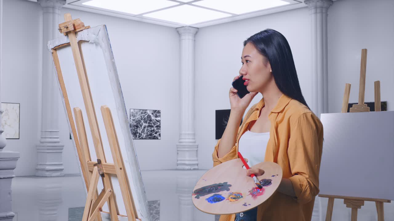 아시아 여성 예술가가가 갤러리에서 오일 페인트와 으로 캔버스에 그림을 그리는 동안 스마트폰으로 이야기합니다.