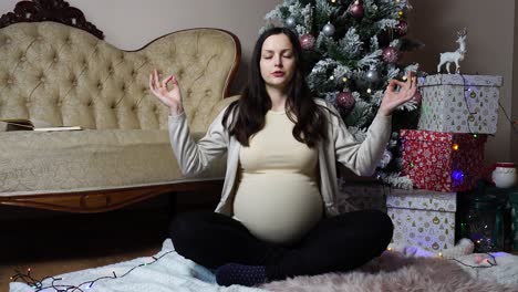 pregnant woman do yoga meditation pose near christmas tree and presents