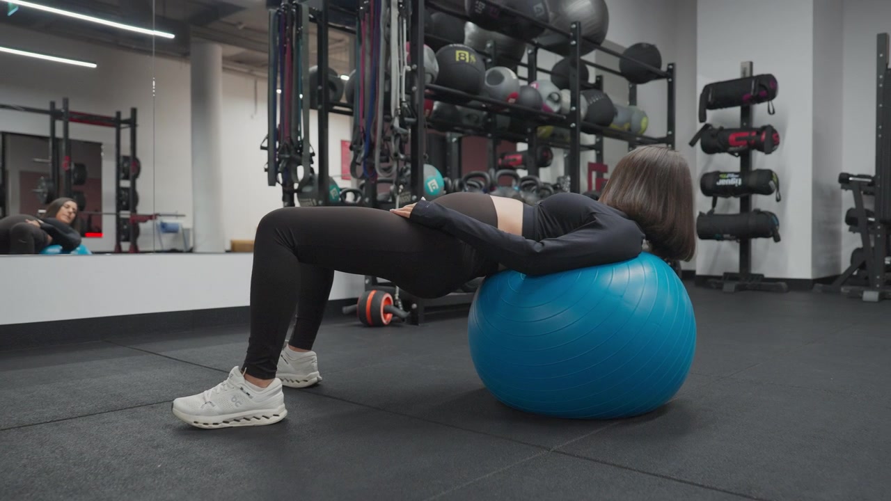 Wide view of a pregnant woman maintaining a supported hip bridge on a blue stability ball during a prenatal gym workout in slow motion.