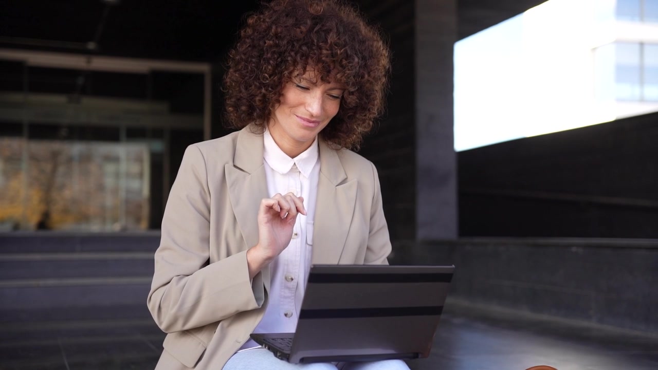 Una donna d'affari sicura di sé sta usando il suo laptop all'aperto in un ambiente vivace, impegnata nel lavoro.