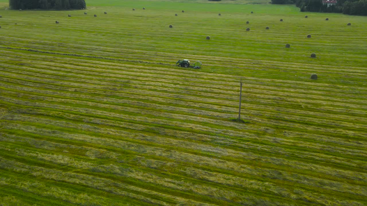 Aerial orbiting around green farm field while rustic hay baler tractor moves across the meadow, gathers and rolling lines of dried and mowed hay, grass crop into round straw bales. Idyllic summer day