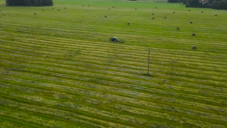aerial orbiting around green farm field while rustic hay baler tractor moves across the meadow, gathers and rolling lines of dried and mowed hay, grass crop into round straw bales. idyllic summer day
