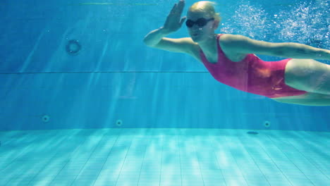 bajo el agua de una hermosa joven rubia sexy con traje de baño rojo de una pieza y gafas, nadando sola y saludando en apnea aguantando la respiración