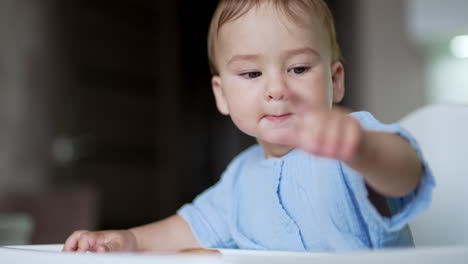 serious thoughtful baby boy sitting at the table. kid playing with food, throwing bagel on the floor and looking at it. close up.