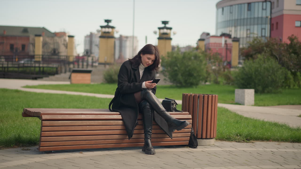 mujer sentada en un banco del parque, usando un teléfono inteligente, vestida con un abrigo negro y botas, rodeada de árboles verdes y exuberantes, disfrutando de una atmósfera al aire libre relajada en un parque urbano, en un entorno pacífico