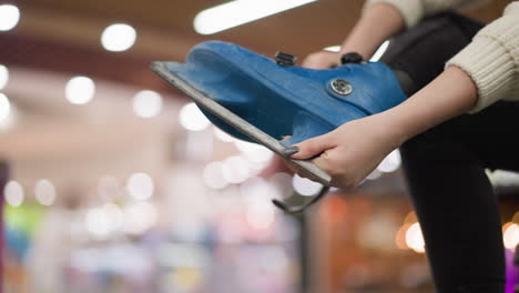 una vista de cerca de una dama con pantalones negros y uñas pintadas quitando un zapato de patinaje sobre hielo azul, con luces borrosas en el fondo, también está en calcetines negros