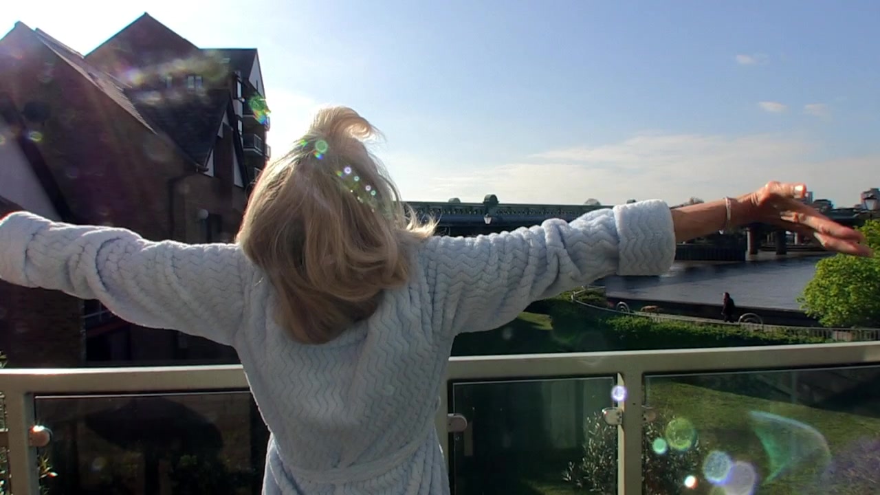 Una donna bionda in vestaglia saluta il giorno dal suo balcone che si affaccia sul fiume Tamigi a Londra