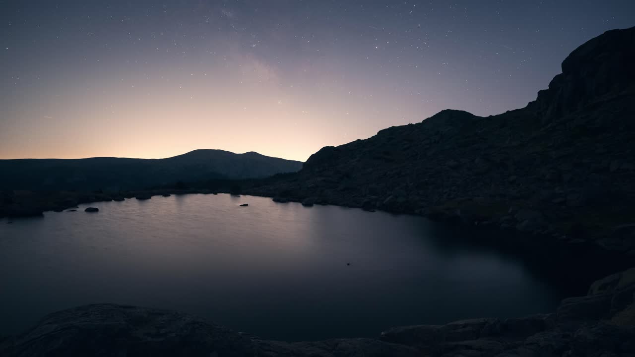 Night timelapse of the Milky Way and stars above Laguna Grande de Peñalara in Sierra de Guadarrama, with light pollution on the horizon