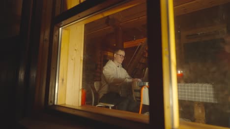 Sneak-peek-through-glass-window-man-sit-near-laptop-in-cozy-wooden-cabin