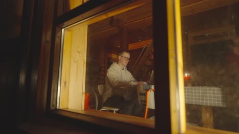 sneak peek through glass window man sit near laptop in cozy wooden cabin