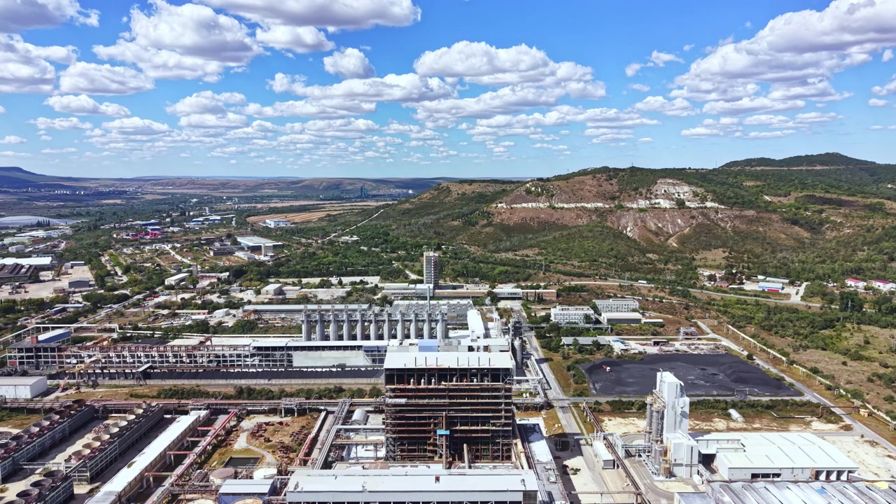 Área industrial com edifícios e colinas ao fundo sob um céu azul com nuvens
