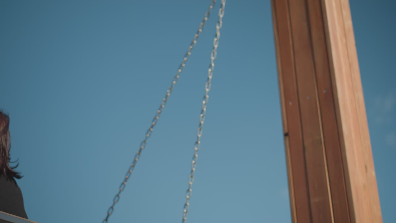 은 파란 하늘 아래 야외에서 흔들리는 여자의 가까운 모습, 약간 미소 짓고, 빛이 그녀의 얼굴에 반사되고 목조 흔들림 사슬이 보입니다.