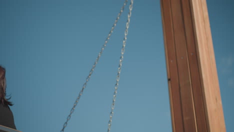 close up of lady swinging outdoors under clear blue sky, smiling slightly, with sunlight reflecting on her face and wooden swing chains in view