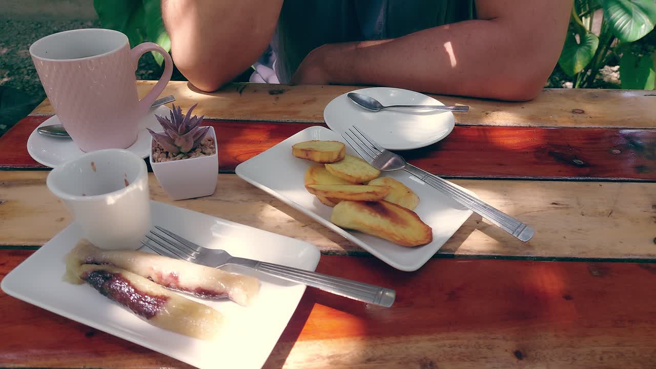 un entorno al aire libre casual con kamote frito, suman y café servido en una mesa de madera y vista parcial de una persona bebiendo café, bocadillos tradicionales filipinos para la merienda