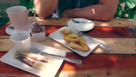 A-casual-outdoor-setting-with-fried-kamote,-suman-and-coffee-served-on-a-wooden-table-and-partial-view-of-a-person-drinking-coffee,-traditional-Filipino-snacks-for-merienda