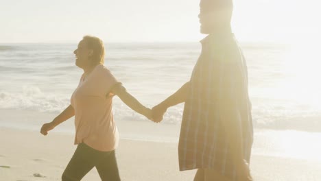 smiling senior african american couple holding hands and walking on sunny beach