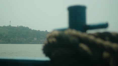 Thick-rope-coiled-around-a-bollard-on-a-boat,-with-blurry-waterscape-and-hills-in-the-background