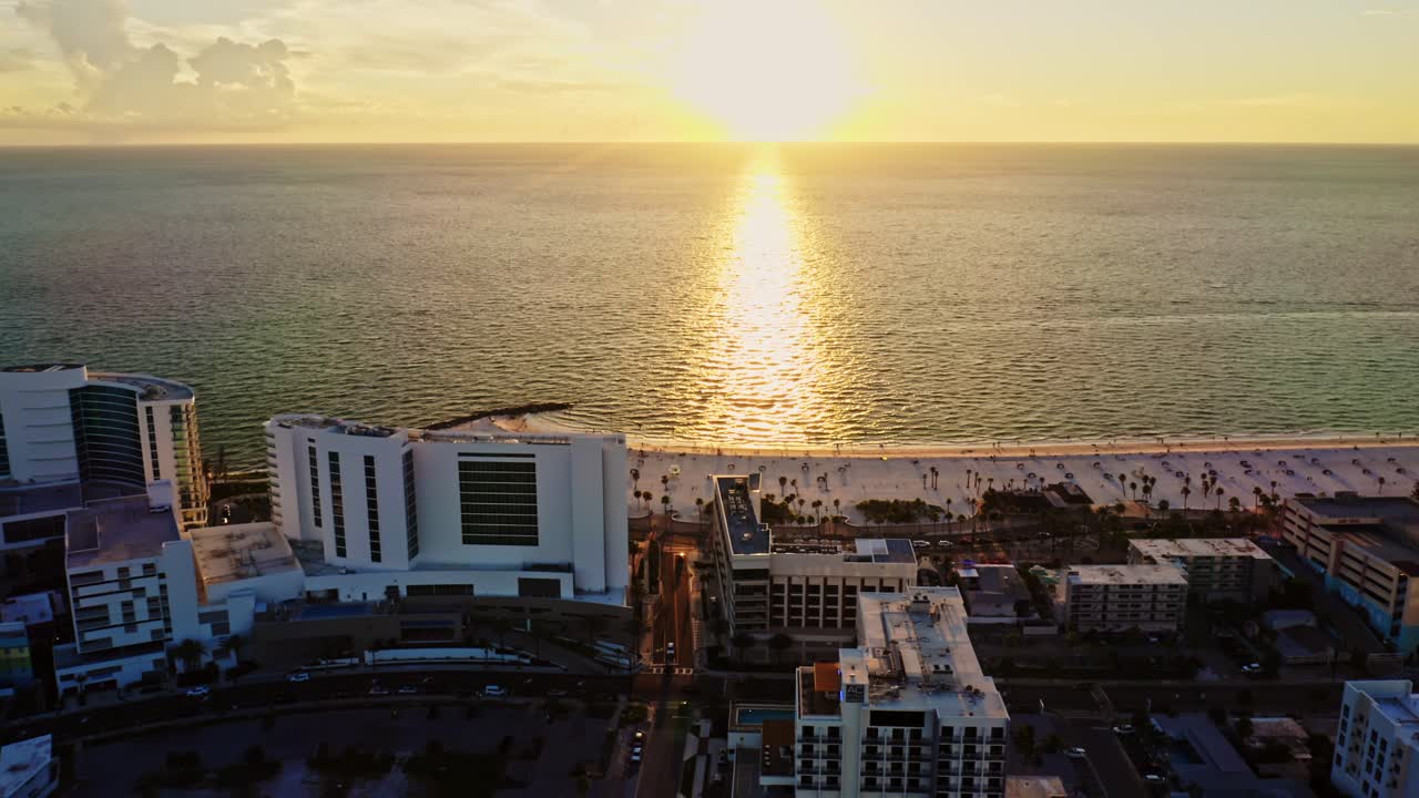 The sun sets over the Gulf of Mexico, casting golden reflections across the water as beachfront resorts and the sandy shoreline of Clearwater Beach glow in evening light