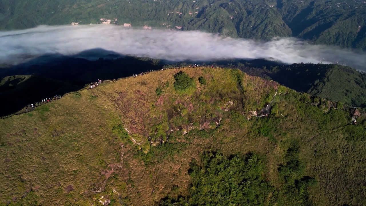 Sti langs vulkanrygg med turgåere som går langs kraterkanten over skyene i gyllent morgenlys, som avslører bratt terreng og dramatisk fjellandskap, luftfoto fra drone