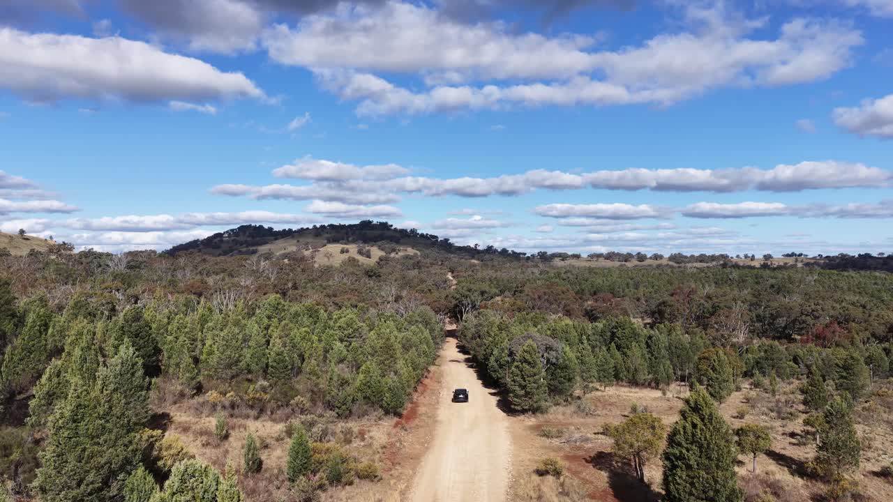 A drone tracks a single vehicle driving along a remote dirt road through bushland near Coonabarabran, New South Wales, under bright daylight and partly cloudy skies
