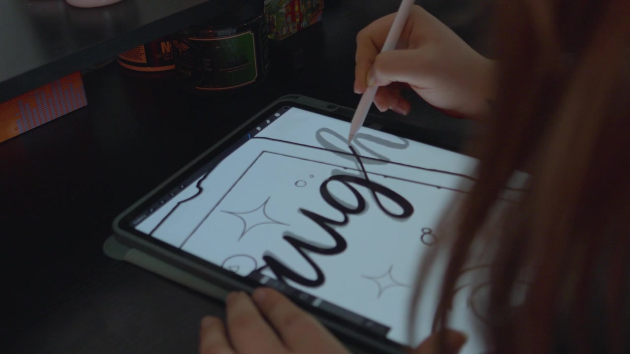 a female tattoo artist with long red hair drawing a design for a tattoo on an tablet as she traces each cursive letter