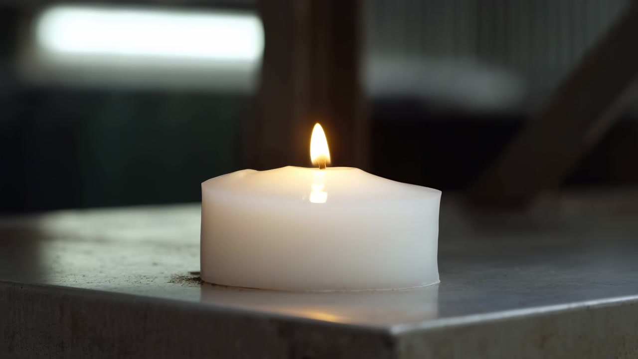Scena in movimento ravvicinata di una singola candela a colonna bianca che brucia con una fiamma gialla costante su un tavolo di pietra opaca, mostrando una pozza di cera fusa, un riflesso morbido, un caldo bagliore, un'atmosfera tranquilla e una superficie texturizzata.