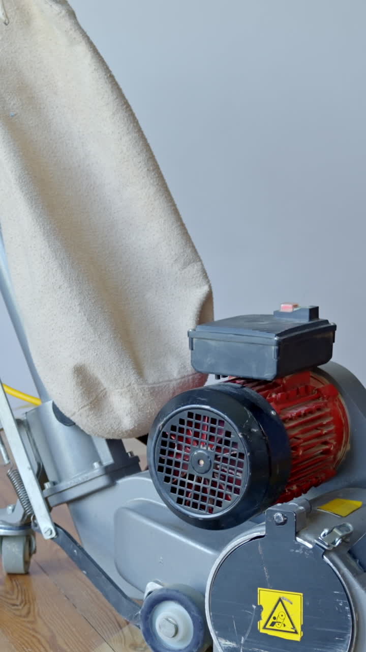 An upward tilting vertical shot reveals heavy-duty floor sanding machine with a red motor and dust bag resting on pine planks during indoor work, camera tilt up, real time