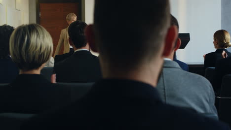 vista posteriore di persone sedute a una conferenza, mentre una donna d'affari entra nella sala conferenze e sale sul podio