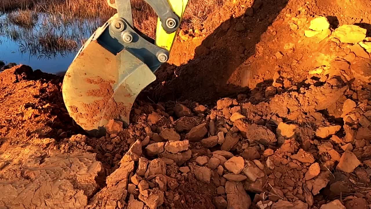 brazo de excavadora excavando tierra roja rica al amanecer en un sitio de construcción, cerca de una fuente de agua, en cámara lenta