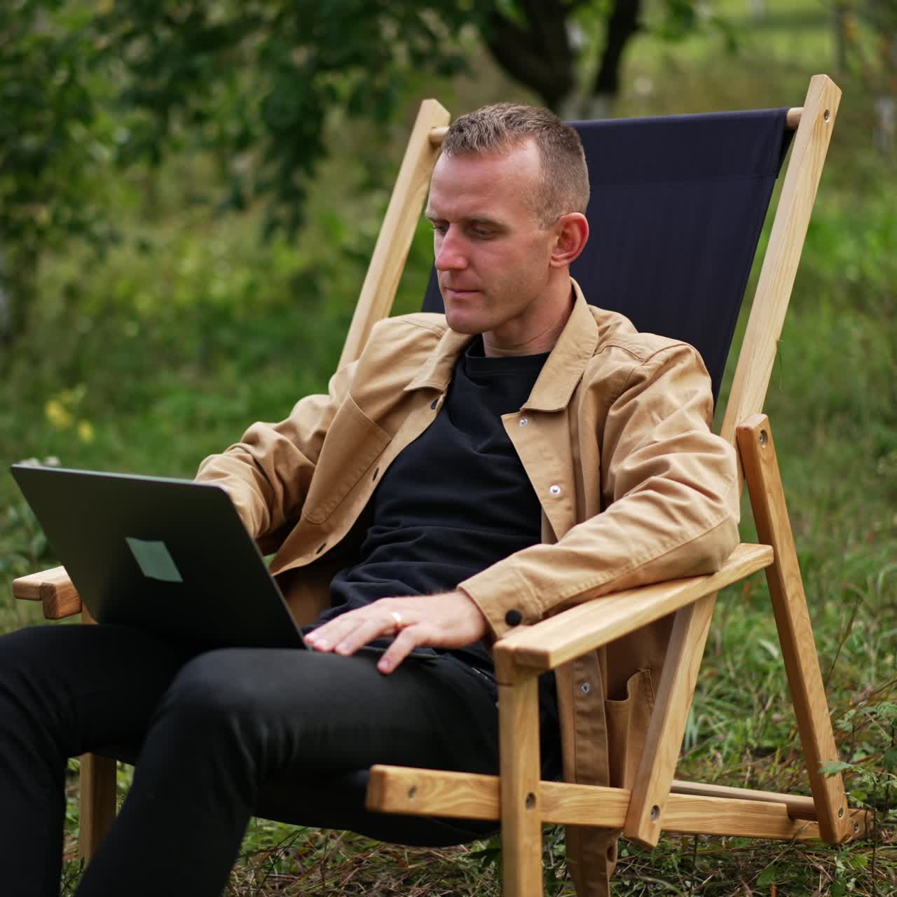 Calm mid-aged man works on his computer outdoors. Freelance IT specialist working remotely from the office. Garden backdrop