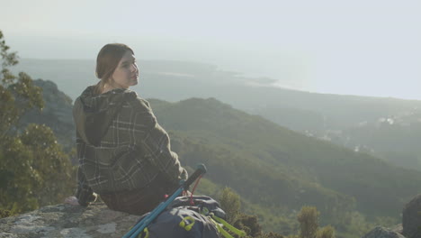 linda excursionista sentada en el borde del acantilado y disfrutando de la vista, luego girando hacia la cámara y sonriendo