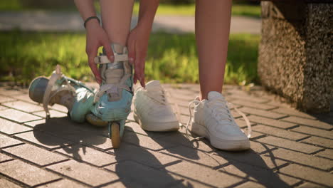 leg view of individual adjusting strap on roller skate of right leg while tapping skate with both hands, wearing black wristband, second roller skate lies by in background, sneakers placed nearby