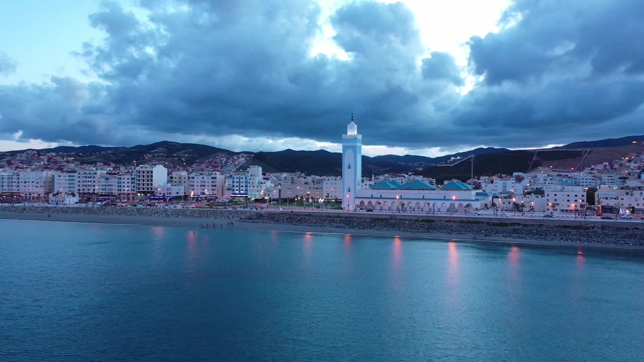 Vista drone all'ora blu sulla tranquilla baia mediterranea di M'diq, con una moschea sul lungomare, luci della città e le montagne del Rif sulla costa del Marocco settentrionale, Nord Africa.